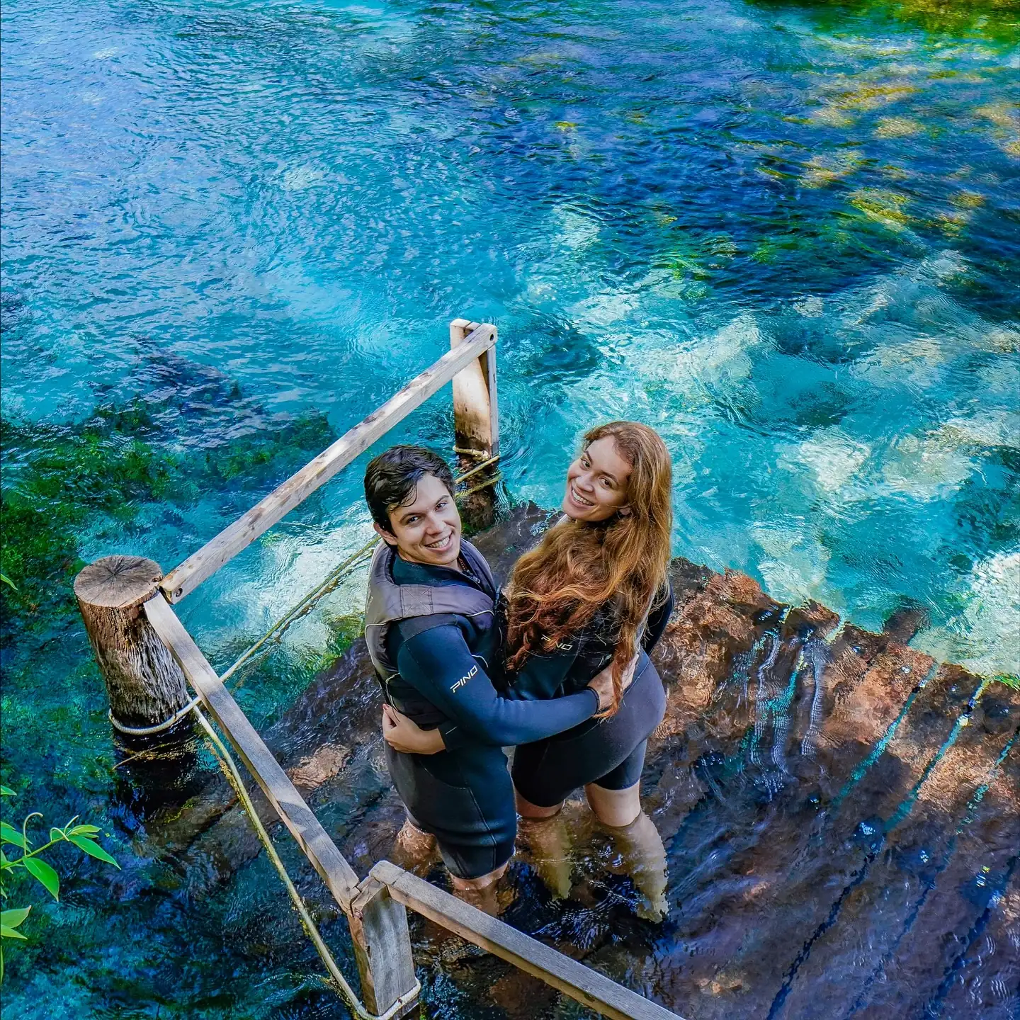 Ju e Ney abraçados num deque sobre a água azul-turquesa de Bonito, olhando para a câmera